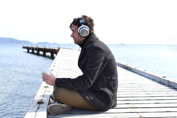Young man listening to music and drinking coffee, sitting alone on pier on a sunny day