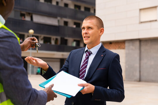 European man in suit reciveing keys to his apartment in new building from African-american man engineer.