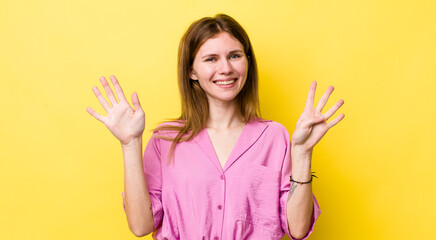 red head pretty woman smiling and looking friendly, showing number nine or ninth with hand forward,...