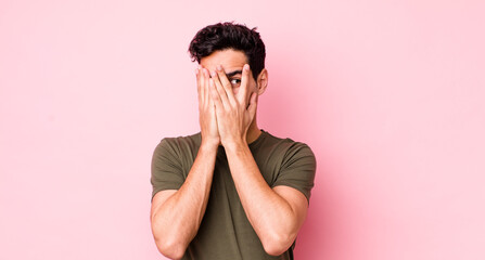 handsome hispanic man covering face with hands, peeking between fingers with surprised expression...