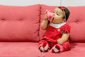 baby latina sitting on a red sofa, chewing on a toy. copy space