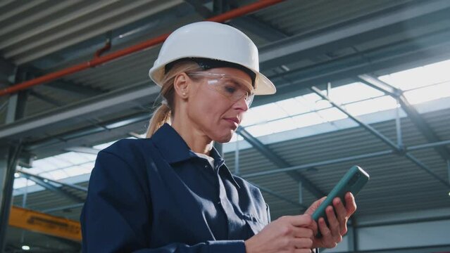 Smiling woman with helmet, uniform and goggles uses a mobile phone in factory at sunlight. In the background details of a metalworking project