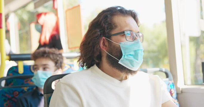 A man with long hair and a thick beard wearing glasses with a mask on his face takes a public transport bus to work