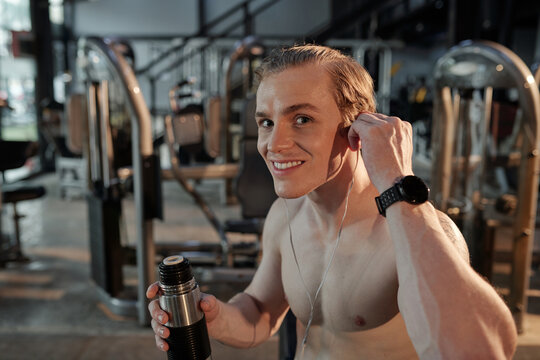 Happy excited young sportsman listening to good music and drinking electrolyte-rich beverage in gym