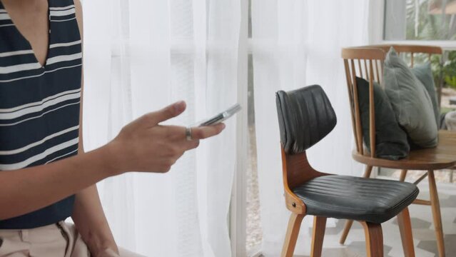 Close up 4K of unrecognised asian young woman who is holding and using her smartphone to connect people by using internet technology in bright and comfortable living room for modern lifestyle.