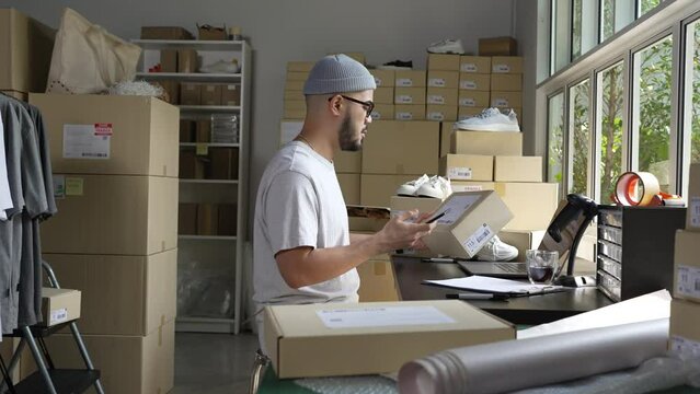 Asian male small business owner using mobile app on smartphone checking parcel box. Warehouse worker, seller holding phone scanning retail dropshipping package postal parcel bar code.