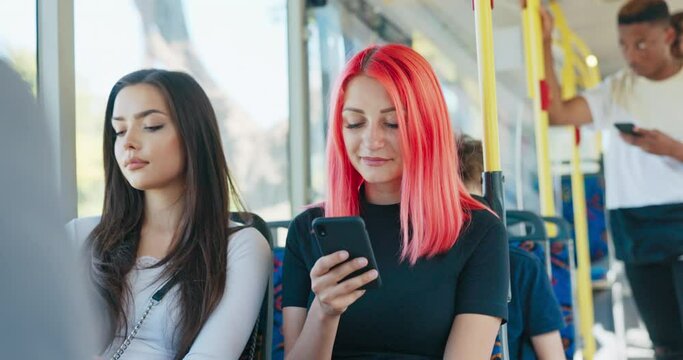 A teenage girl with pink hair sits in a chair on a public transit bus next to another woman, browsing social media on her phone, replying to a friend's messages, wondering