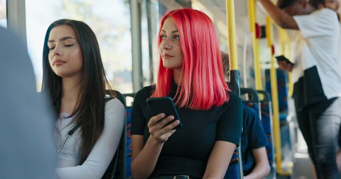 A teenage girl with pink hair sits in a chair on a public transit bus next to another woman, browsing social media on her phone, replying to a friend's messages, wondering, smiling, laughing