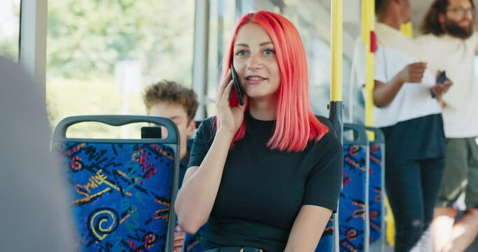 A beautiful smiling girl with pink hair is riding a bus to meet a friend. The woman is sitting on a chair talking on the phone, laughing, enjoying the journey by public transport.