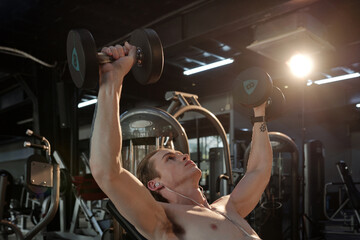 Fit young man listening to music in earbuds when doing dumbbell press on bench