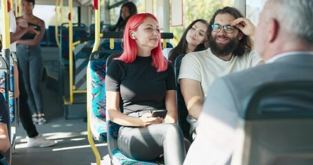 Girl with pink hair rides public transport bus, talks to friend sitting next to her, woman holds smartphone in hands, has wireless headphones in ears - Powered by Adobe