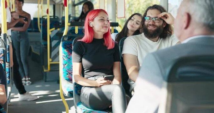 Girl with pink hair rides public transport bus, talks to friend sitting next to her, woman holds smartphone in hands, has wireless headphones in ears