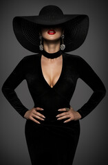 Fototapeta na wymiar Sexy Fashion Woman in Big Hat and Black Dress. Beauty Model with Curve Body Shapes in Seductive Bodycon Gown. Mysterious Elegant Lady over Dark Gray Background. Women hidden Face and Red Lips