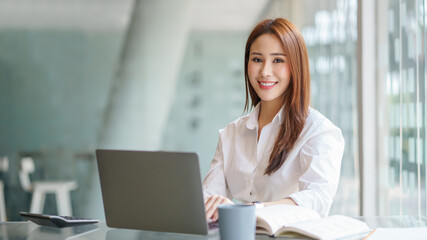 Beautiful young Asian businesswoman sitting working with a laptop at the office. Looking at camera.