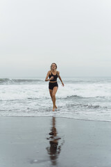 Fototapeta premium A barefoot young woman with a slender body runs on the sea surf by the water pool to keep fit and burn fat. Beach background with blue sky. Women's fitness, jogging, sports activities 