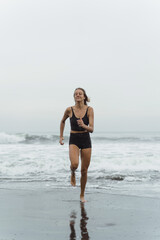 Fototapeta premium A barefoot young woman with a slender body runs on the sea surf by the water pool to keep fit and burn fat. Beach background with blue sky. Women's fitness, jogging, sports activities 