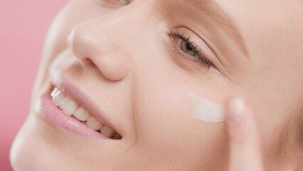Extreme close-up of young Caucasian woman with perfect skin applies face care cream to her cheek under her eye smiling wide on pink background | Face and eye cream commercial concept