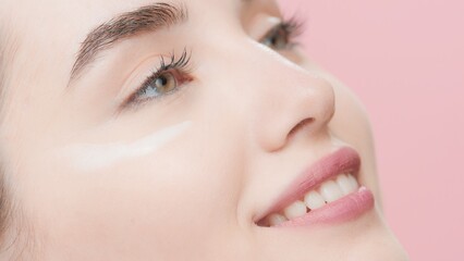 Extreme close-up of young beautiful fit Caucasian skin care woman with cream smear under her eye smiling wide against pink background | Face cream commercial concept