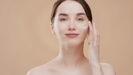 Naklejka premium Young positive Caucasian woman with brown hair and bare shoulders applies face cream on her cheek against beige background with water ripples | Face cream commercial concept