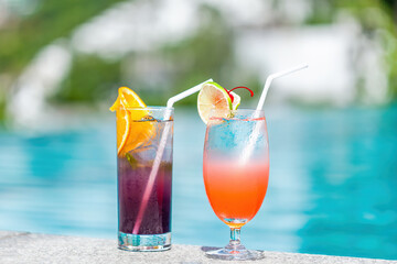 Vacation, summer, holiday, luxury resort concept. Two glasses with tropical cold cocktails with ice, plastic straw and slices of lime and orange near blue swimming pool in hot summer day