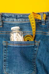 yellow measuring tape and a glass jar with pills are in the back pocket of blue jeans on a yellow background., vertical photo.