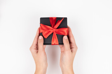 Close up of female hands holding black gift box with red silk ribbon over white background