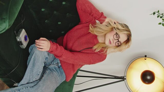 Vertical video, Blonde woman wearing glasses, jeans and a red sweatshirt, listens to music from a walkman with headphones. She sits on a green velvet armchair surrounded by a plant and a vintage lamp.