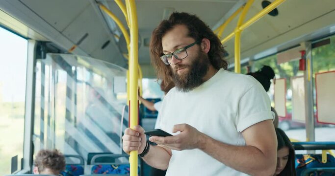 Hipster man with long hair and beard wearing glasses travels home in public transportation vehicle, guy holds onto handrail, scrolls social media on phone