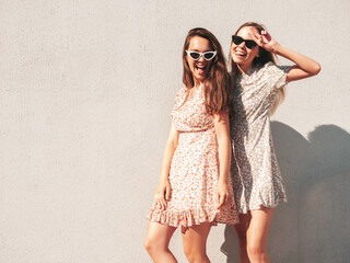 Two young beautiful smiling hipster female in trendy summer dresses. carefree women posing on the street background near white wall. Positive pure models having fun at sunset, hugging