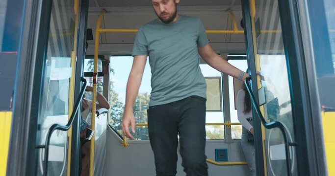 The Doors Of A Public Transportation Vehicle Open After Coming To A Stop. A Man Gets Off The Bus Followed By A Student And A Woman Of African-American Descent, People From The Street Get On The Bus