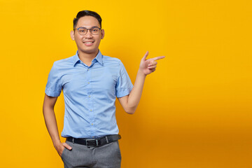 portrait of smiling young asian man in glasses pointing finger away at copy space isolated on yellow background. businessman and entrepreneur concept