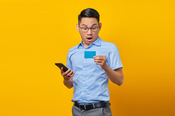 Portrait of amazed young asian man Asian in glasses holding mobile phone and credit card isolated on yellow background. businessman and entrepreneur concept