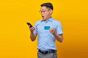 Portrait of amazed young asian man Asian in glasses holding mobile phone and credit card isolated on yellow background. businessman and entrepreneur concept