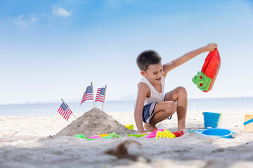 A cute boy playing toy on the beach with a pile of sand with the American flag embroidered near.