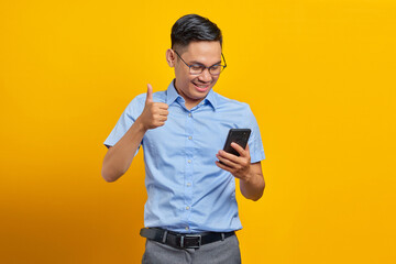 Smiling young handsome business man in classic blue shirt using mobile phone and showing thumbs up gesture isolated on yellow background. Achieve career wealth business concept