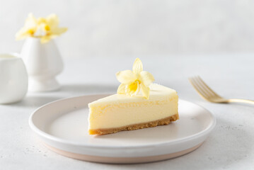 Obraz na płótnie Canvas Classic New York cheesecake with fresh vanilla flower on white concrete background, side view. A piece of Vanilla cheesecake on a white plate. Confectionery menu, recipe. Close up.