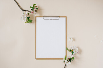 Clipboard tablet pad with blank copy space and white apple flowers on beige backdrop. Flat lay, top view template. Female office desk