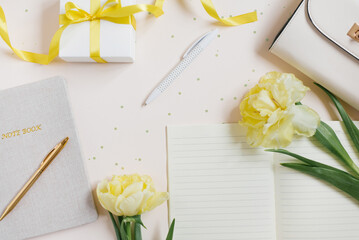 Workspace of a female blogger. Yellow flowers of spring tulips, notebooks with a pen, womens bag, gift with a yellow ribbon on a light background