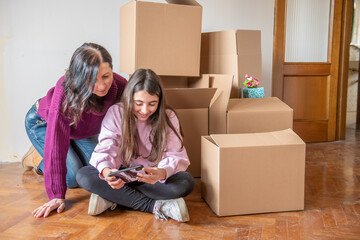 Fototapeta na wymiar Moving to a new home concept. Young girl and her mother seated near cardboard boxes along the parquet floor playing with smartphone.