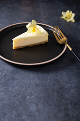 Classic New York cheesecake with fresh vanilla flower on dark concrete background, side view. A piece of Vanilla cheesecake on a black plate. Confectionery menu, recipe. Copy space, vertical