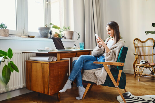 Smiling freelancer using mobile phone sitting on chair at home - Powered by Adobe