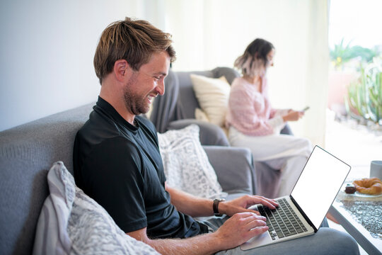 Happy freelancer working on laptop with woman using smart phone in living room