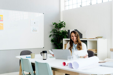 Smiling businesswoman holding paper sitting with laptop at desk