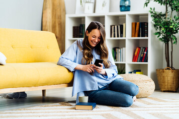 Happy woman using smart phone sitting on carpet by sofa at home