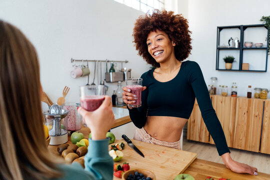 Happy young woman holding glass of smoothie talking with friend in kitchen