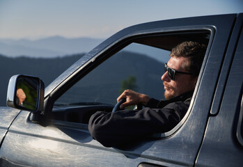 Close up of male in sunglasses driving black car looking to the left at mountain landscape while riding in mountain spaces in sunshine afternoon.