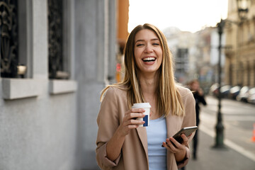 Portrait of happy business woman with coffee and mobile phone on her way to work on city street.