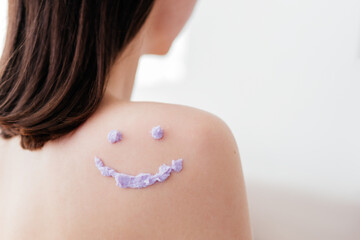 Smiley face made of purple cream scrub on shoulder of unrecognizable woman indoors. Skincare concept