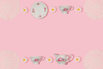 Spring creative frame with vintage tea cups, saucers, embroidery lace placemat and flower heads on pastel pink background. 80s, 90s retro romantic aesthetic summer concept. Fashion tea party idea.