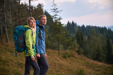 Fototapeta na wymiar Portrait of man and woman with hiking equipment holding hands together, preparing start hike in the mountains. Green grass and coniferous forest on the background. Concept of hiking, relationships.